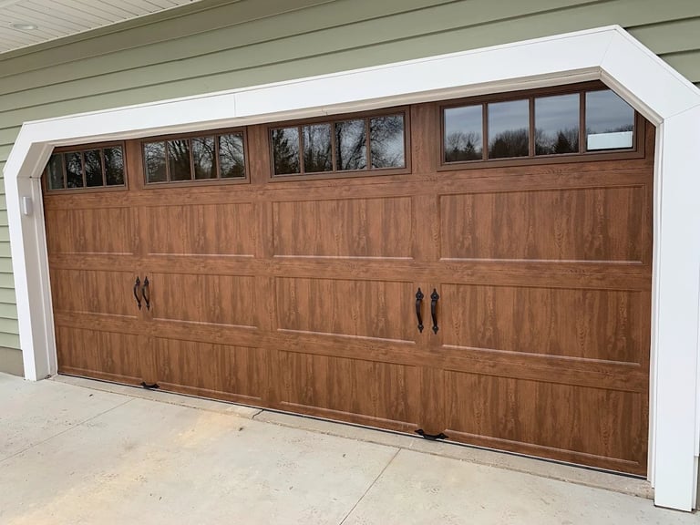 Brown wooden garage door with windows on a white-trimmed garage attached to a house with gray siding